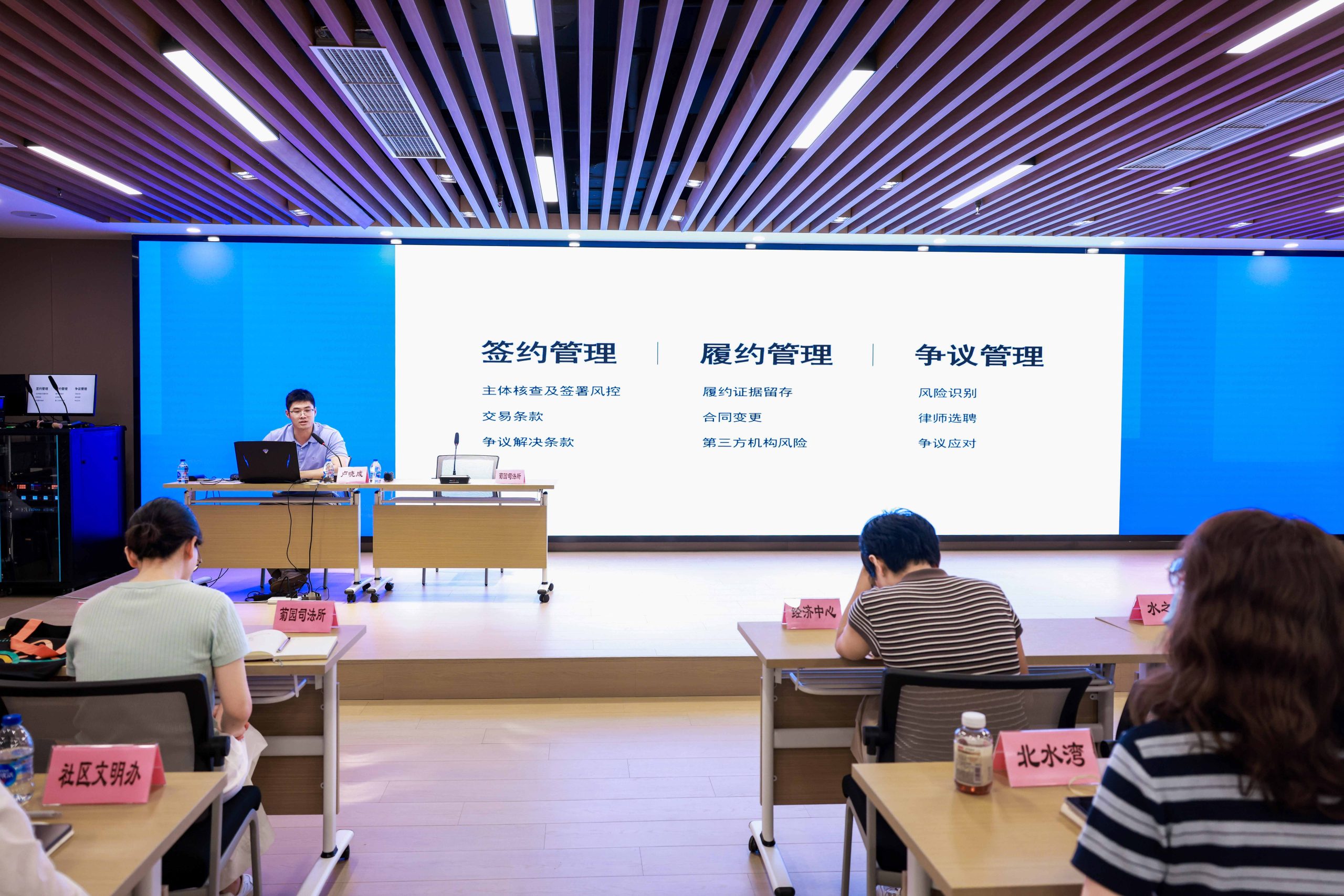 讲座现场,讲师在台上发言,大屏幕展示签约管理、履约管理、争议管理三大模块,台下与会者聆听