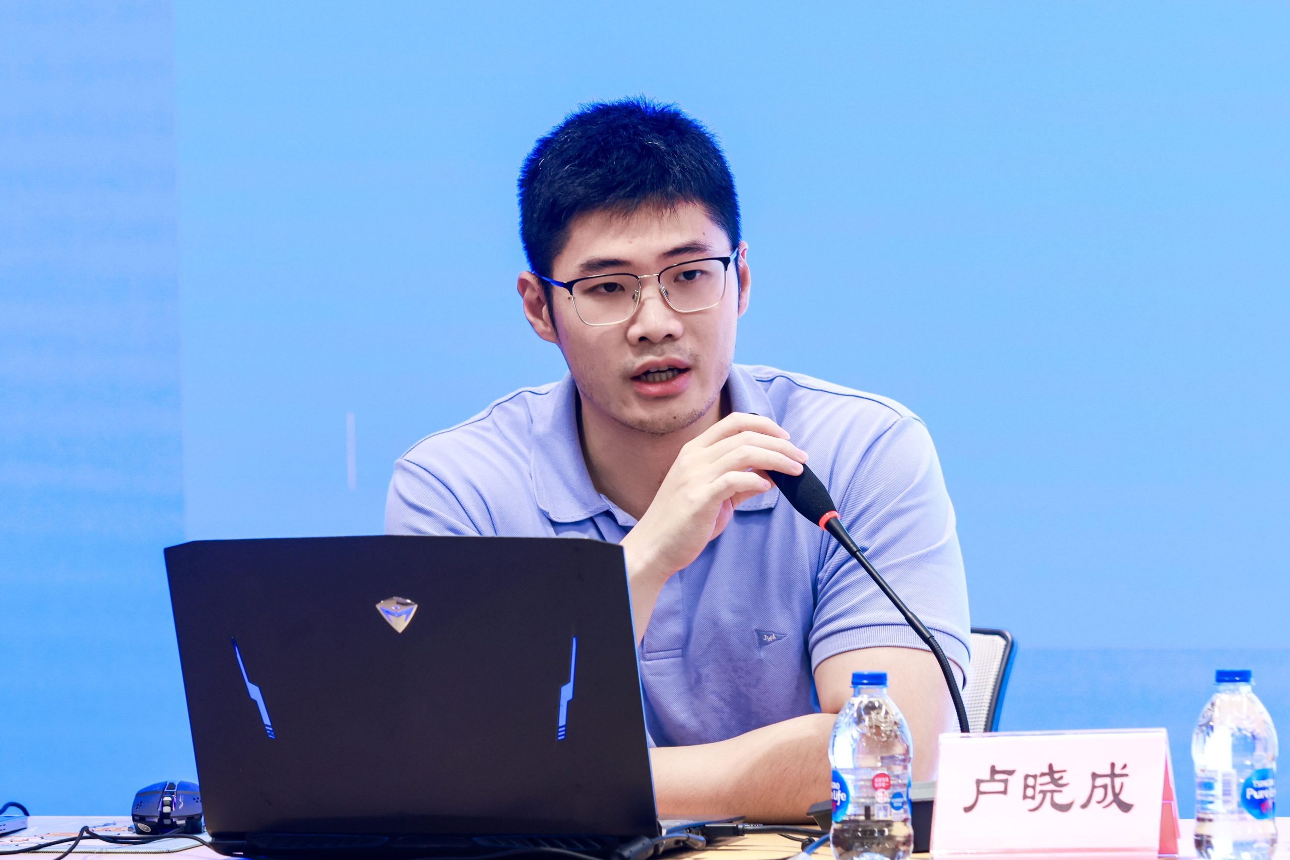 卢晓成戴着眼镜,在蓝色背景前对着麦克风发言,面前放有笔记本电脑